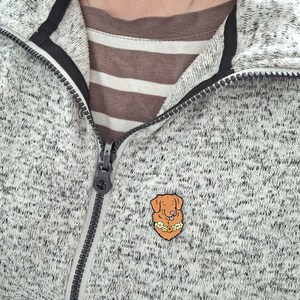A Toller pin badge shown being worn on a white mottled zip-up jumper.