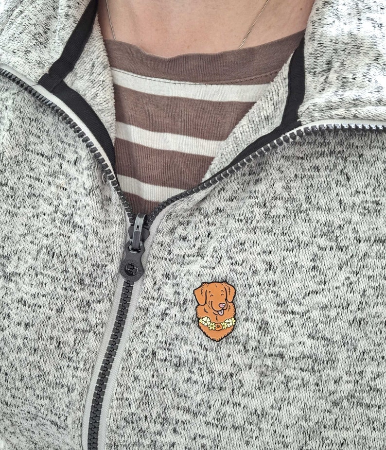A Toller pin badge shown being worn on a white mottled zip-up jumper.