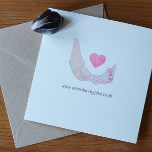 Puede incluir: Una tarjeta de felicitaci&oacute;n cuadrada de color blanco con una ilustraci&oacute;n impresa de una mano grande sosteniendo una mano peque&ntilde;a con un coraz&oacute;n rosa encima. El texto www.animalartbyjess.co.uk est&aacute; impreso en gris debajo de la ilustraci&oacute;n. Una peque&ntilde;a figura de conejo marr&oacute;n se encuentra en la esquina.