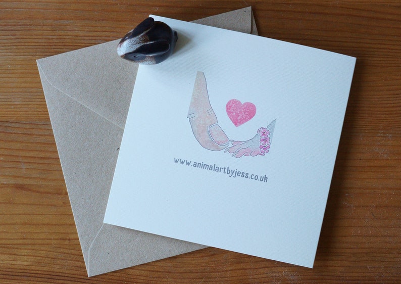 Puede incluir: Una tarjeta de felicitaci&oacute;n cuadrada de color blanco con una ilustraci&oacute;n impresa de una mano grande sosteniendo una mano peque&ntilde;a con un coraz&oacute;n rosa encima. El texto www.animalartbyjess.co.uk est&aacute; impreso en gris debajo de la ilustraci&oacute;n. Una peque&ntilde;a figura de conejo marr&oacute;n se encuentra en la esquina.