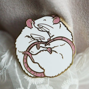 An enamel pin showing two white rats hugging, with gold coloured plating.