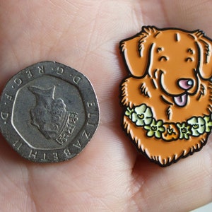 A hand holding a 20p piece and a Nova Scotia Duck Tolling Retriever enamel pin badge. The Toller pin is about the same width as the coin, and taller by approximately fifty percent.
