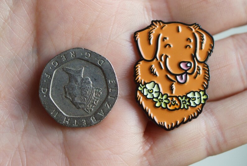 A hand holding a 20p piece and a Nova Scotia Duck Tolling Retriever enamel pin badge. The Toller pin is about the same width as the coin, and taller by approximately fifty percent.