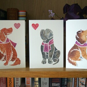 May include: Three greeting cards with watercolor illustrations of dogs looking up at a pink heart. The dogs are wearing pink scarves. The cards are on a shelf with books behind them.