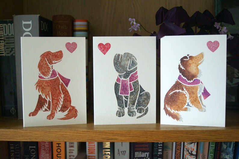 May include: Three greeting cards with watercolor illustrations of dogs looking up at a pink heart. The dogs are wearing pink scarves. The cards are on a shelf with books behind them.