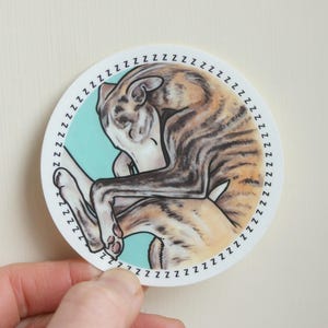 A hand holding up a sleeping Whippet dog sticker against a beige background