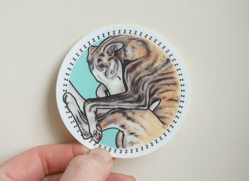 A hand holding up a sleeping Whippet dog sticker against a beige background