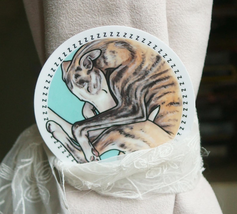 A vinyl sticker of a sleeping Whippet dog, curled up with a blue background and zzzzz all the way around the outside. The sticker is circular.