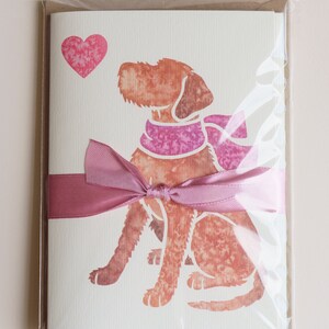 May include: A pack of five blank greeting cards featuring a watercolor illustration of a brown dog wearing a pink scarf and looking at a pink heart. The cards are tied together with a pink ribbon and packaged in a clear plastic sleeve.
