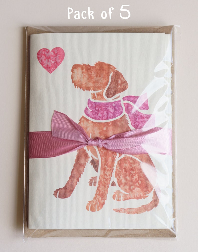 May include: A pack of five blank greeting cards featuring a watercolor illustration of a brown dog wearing a pink scarf and looking at a pink heart. The cards are tied together with a pink ribbon and packaged in a clear plastic sleeve.