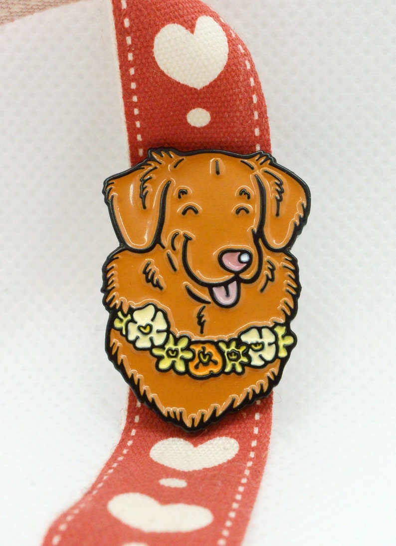 A Toller enamel pin badge shown on a white background, with a red and white heart pattern ribbon. The pin shows the head and neck of a happy smiling dog, with a yellow flower garland. The enamel is shiny and recessed (soft enamel).