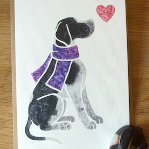 May include: A black and white dog wearing a purple scarf sits looking at a red heart. The dog is sitting on a white background.