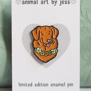 A Toller pin badge shown on a white backing card. The cardstock is textured and there is a light grey heart shape in the centre where the pin is attached. There is text with the artist name and website address.
