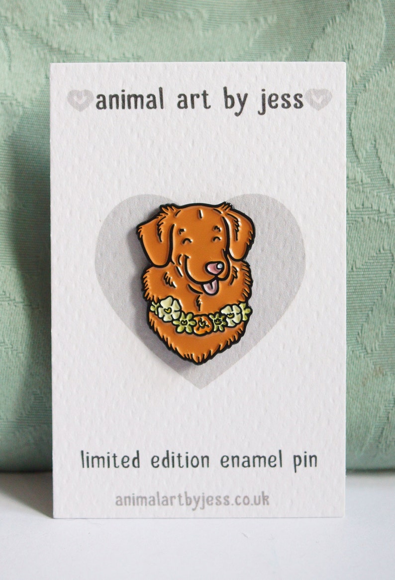 A Toller pin badge shown on a white backing card. The cardstock is textured and there is a light grey heart shape in the centre where the pin is attached. There is text with the artist name and website address.