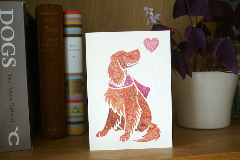 May include: A watercolor illustration of a dog wearing a pink scarf, looking up at a pink heart. The illustration is on a white card.