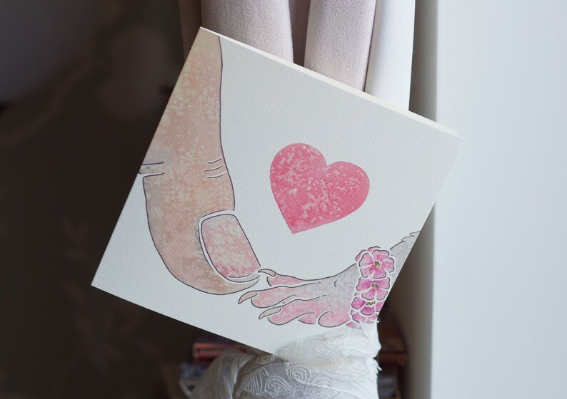 Puede incluir: Un lienzo cuadrado con una ilustraci&oacute;n en acuarela de un pulgar humano grande tocando la mano de una peque&ntilde;a criatura con garras. Un coraz&oacute;n rosa texturizado se sit&uacute;a entre ambas figuras y la criatura lleva una pulsera de flores rosas.