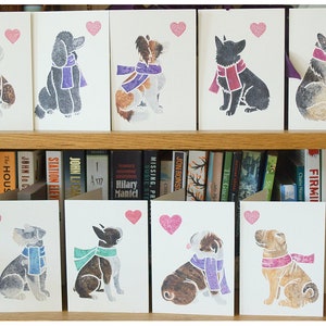 May include: Ten greeting cards featuring different dog breeds wearing scarves and a heart. The cards are white with a pink heart in the top right corner. The dogs are all in a sitting position and are facing the viewer. The cards are arranged in two rows of five on a bookshelf.