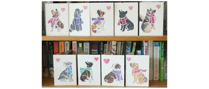 May include: Ten greeting cards featuring different dog breeds wearing scarves and a heart. The cards are white with a pink heart in the top right corner. The dogs are all in a sitting position and are facing the viewer. The cards are arranged in two rows of five on a bookshelf.