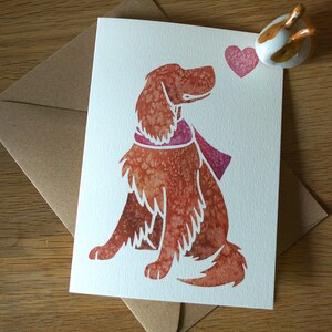 May include: A watercolor illustration of a brown dog with a pink scarf looking at a pink heart. The dog is sitting on a white card with a brown envelope behind it.