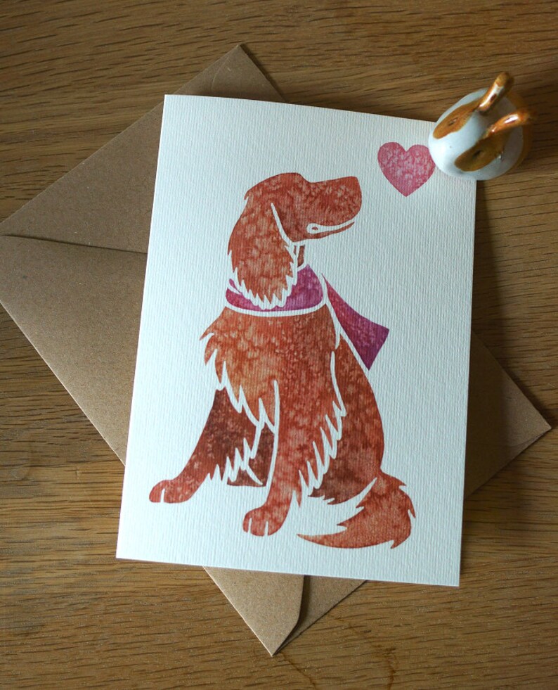 May include: A watercolor illustration of a brown dog with a pink scarf looking at a pink heart. The dog is sitting on a white card with a brown envelope behind it.