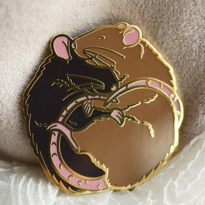 An enamel pin featuring two sleeping rats cuddled up in a circle. The left hand side rat is black and the right hand side is brown/agouti.