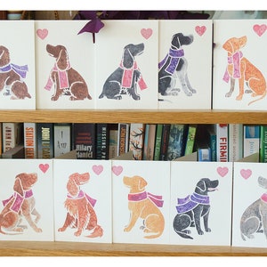 May include: A collection of 16 greeting cards featuring illustrated dogs wearing scarves. The cards are arranged in two rows on a shelf with books behind them. Each card has a pink heart in the top right corner.