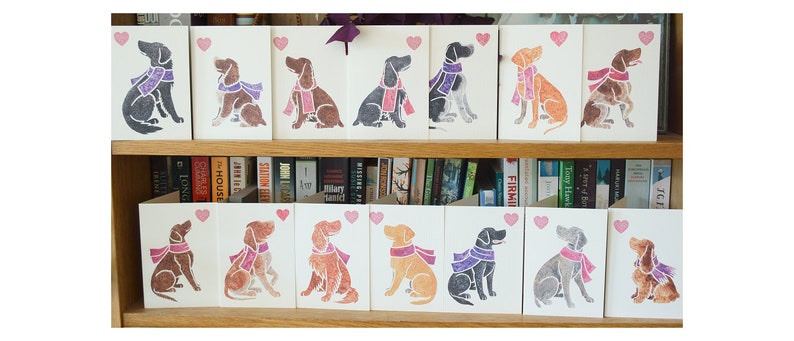 May include: A collection of 16 greeting cards featuring illustrated dogs wearing scarves. The cards are arranged in two rows on a shelf with books behind them. Each card has a pink heart in the top right corner.
