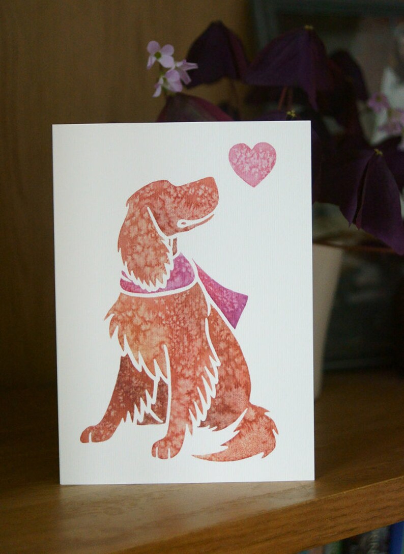 May include: A watercolor illustration of a dog with a pink scarf looking up at a pink heart. The dog is sitting and has a brown and pink color scheme.