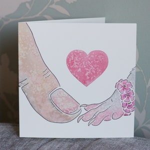 Puede incluir: Una tarjeta de felicitaci&oacute;n cuadrada con una ilustraci&oacute;n en acuarela de un dedo humano tocando la mano con garras de una criatura. Un coraz&oacute;n rosa texturizado est&aacute; centrado sobre las manos. La mu&ntilde;eca de la criatura lleva una pulsera de peque&ntilde;as flores rosas.