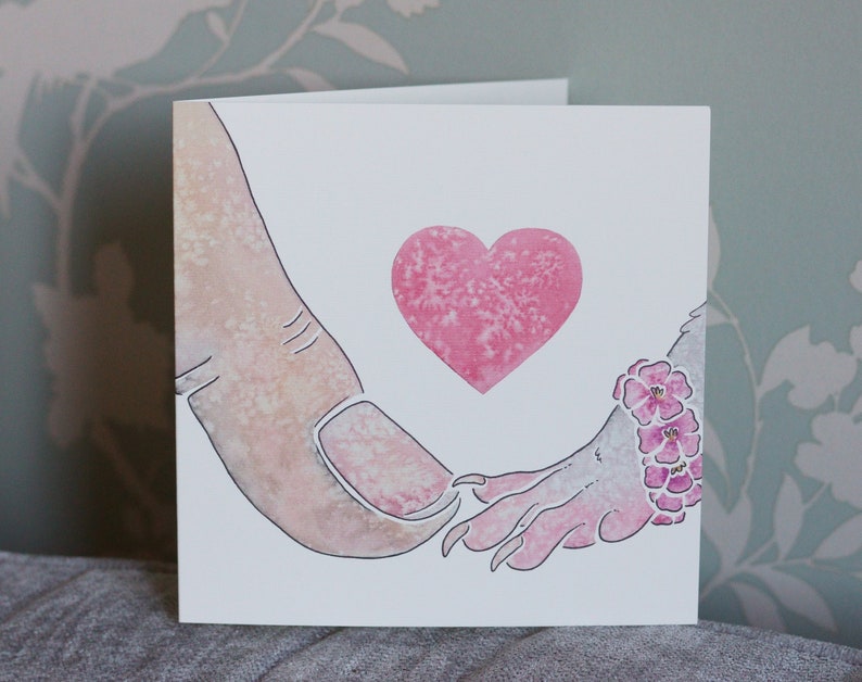 Puede incluir: Una tarjeta de felicitaci&oacute;n cuadrada con una ilustraci&oacute;n en acuarela de un dedo humano tocando la mano con garras de una criatura. Un coraz&oacute;n rosa texturizado est&aacute; centrado sobre las manos. La mu&ntilde;eca de la criatura lleva una pulsera de peque&ntilde;as flores rosas.