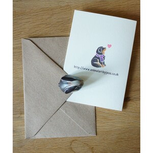 May include: A greeting card with a black and brown dog wearing a purple scarf. The dog is looking up at a pink heart. The text on the card reads "http://www.animalartbyjess.co.uk".