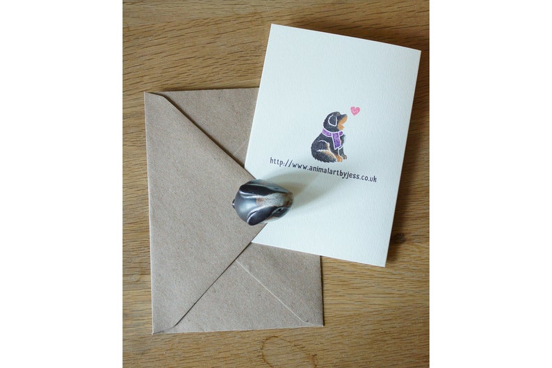 May include: A greeting card with a black and brown dog wearing a purple scarf. The dog is looking up at a pink heart. The text on the card reads "http://www.animalartbyjess.co.uk".