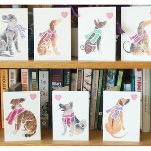 May include: Set of eight greeting cards featuring watercolor illustrations of dogs wearing scarves. Each card has a pink heart in the top right corner.