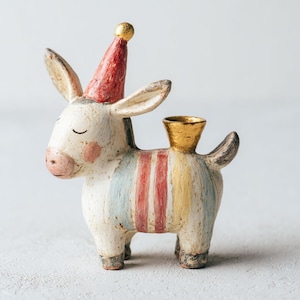 Puede incluir: Figura de burro de cerámica con un gorro de fiesta rojo y detalles dorados. El burro tiene un cuerpo blanco con rayas de colores y un portavelas dorado en la espalda. Un objeto decorativo.