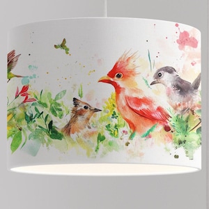 Puede incluir: Una pantalla de lámpara blanca con un diseño de acuarela de pájaros y flores. Los pájaros son rojos, naranjas, marrones y grises. Las flores son rosas, amarillas y verdes.