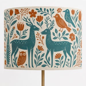 May include: A cylindrical lampshade with a nature-inspired design. It features teal deer, birds, and an owl, alongside orange flowers and green foliage, set against a cream background. The lampshade is supported by a gold-coloured metal stand.