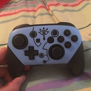 May include: A blue Nintendo Switch controller with black buttons and a stylized eye design. The controller has a black and white design on the front.