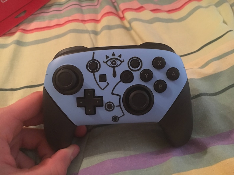 May include: A blue Nintendo Switch controller with black buttons and a stylized eye design. The controller has a black and white design on the front.