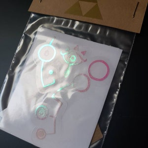 May include: A set of holographic stickers featuring various designs from the Legend of Zelda video game series. The stickers are iridescent and have a pink and green color scheme.