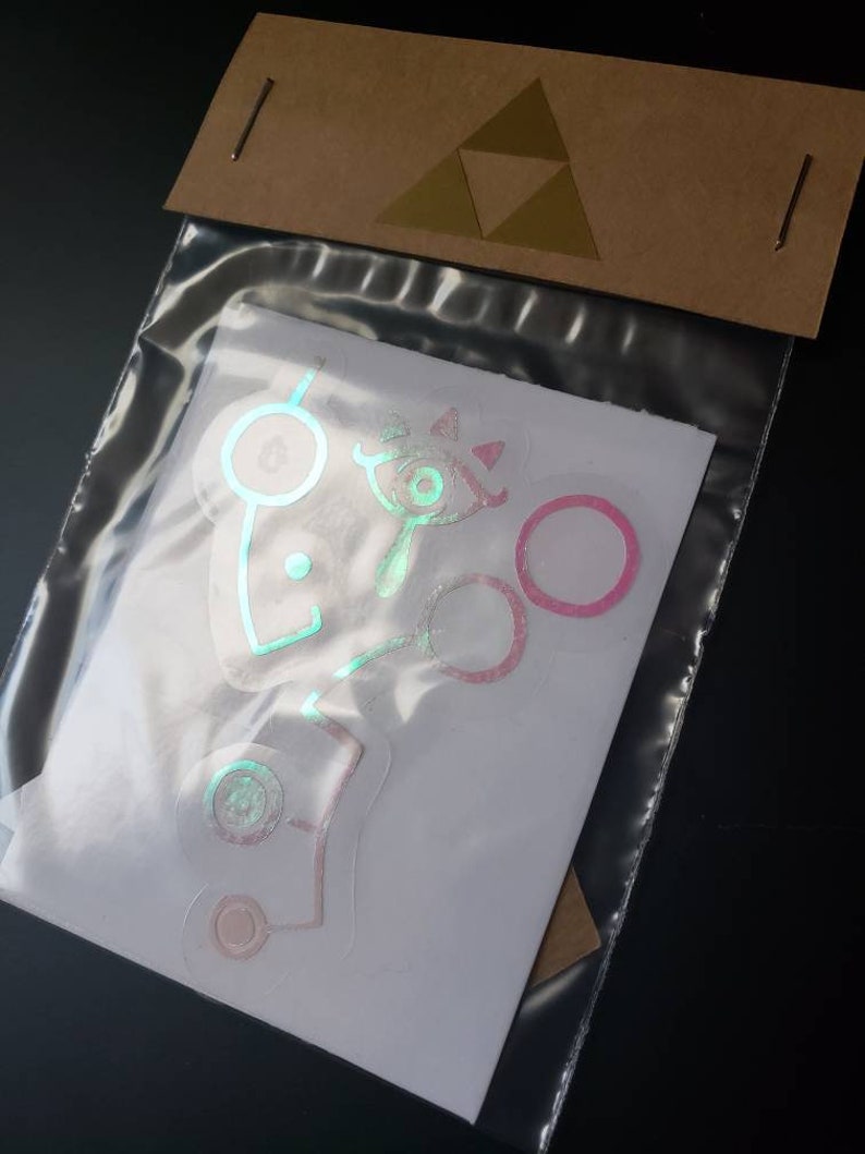May include: A set of holographic stickers featuring various designs from the Legend of Zelda video game series. The stickers are iridescent and have a pink and green color scheme.