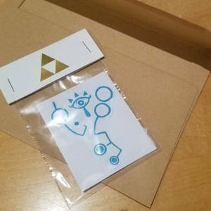 May include: A white envelope with a gold triangle logo and a small, clear plastic bag containing a blue and white sticker with a stylized eye and geometric shapes.