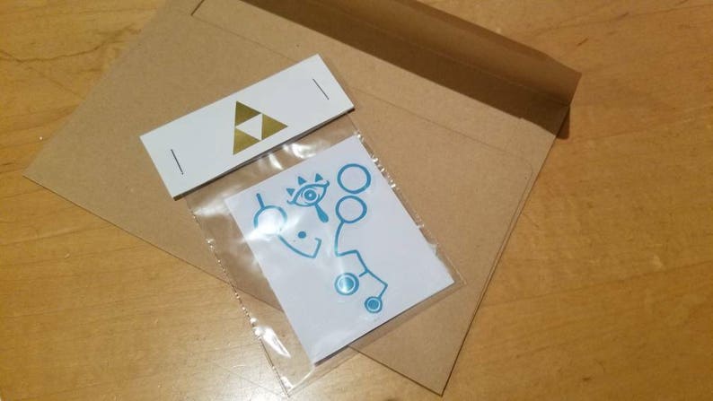 May include: A white envelope with a gold triangle logo and a small, clear plastic bag containing a blue and white sticker with a stylized eye and geometric shapes.