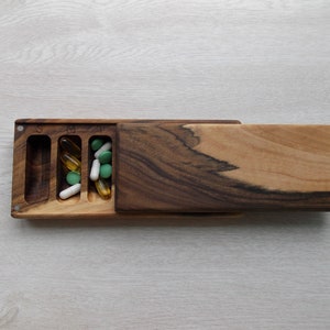 May include: A wooden pill organizer with a sliding lid. The organizer is divided into three compartments labeled S, M, and T, each containing different colored pills and capsules. The wood grain is visible.