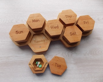 May include: Wooden pill organizer with seven compartments labeled with days of the week: Sun, Mon, Tue, Wed, Thu, Fri, Sat. The organizer is shaped like a honeycomb. A separate compartment is open and contains several pills.