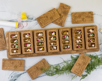 Puede incluir: Un organizador de pastillas de madera con siete compartimentos etiquetados del domingo al sábado. Cada compartimento está lleno de varias pastillas y cápsulas. El organizador está hecho de madera marrón claro y tiene un acabado de madera natural.