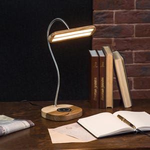 Puede incluir: Una lámpara de escritorio de madera con un cabezal de luz rectangular y un cuello flexible de color plateado. La lámpara está sobre una base de madera. El escritorio también tiene libros, un cuaderno, un bolígrafo y un periódico enrollado.