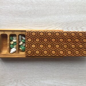 May include: Wooden pill box with a honeycomb pattern lid. The box has seven compartments labelled "S", "M", "T", "W", "Th", "F", and "Sa".