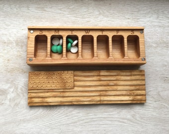 May include: Wooden pill box with seven compartments labeled S, M, T, W, T, F, S. The box is open and contains green and white pills. The box is sitting on a wooden surface.