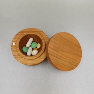 Pode incluir: Uma caixa de pílulas de madeira com tampa articulada, aberta para revelar uma variedade de pílulas e cápsulas em verde, branco e amarelo.