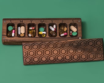 May include: A wooden pill box with a honeycomb pattern on the lid. The box is open and has seven compartments labeled S, M, T, W, T, F, and S. Each compartment contains various pills and capsules.
